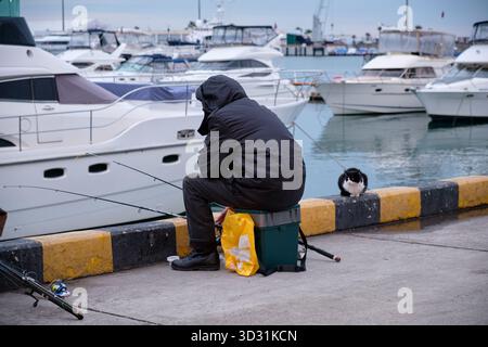 Er sah fisher, der auf dem Pier der Grand Marina saß, mit einer Katze, die auf den Fang wartete. Seehafen von Sotschi. Region Krasnodar, Russland Stockfoto