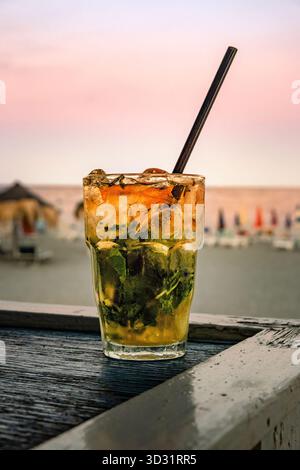 Ein erfrischender Mojito-Cocktail mit Pfefferminze und Eis sitzt bei Sonnenuntergang auf einer hölzernen Strandbar, während das Meer im Hintergrund verschwimmt. Stockfoto