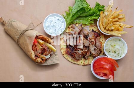 Griechische Gyros-Platte und -Wrap mit Pommes, Tzatziki, Salat, Zwiebeln und Tomaten auf braunem Papierhintergrund bei natürlichem Tageslicht Stockfoto