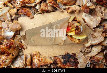 Nahaufnahme eines frisch gewickelten Huhn-Gyros, umgeben von gegrillten Fleischstücken auf einer rustikalen Holzoberfläche, die in natürlichem Tageslicht mit warmen Tönen erfasst wird Stockfoto