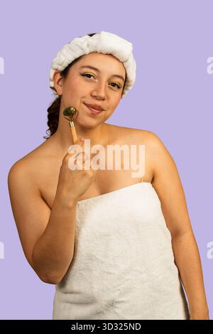 Frau im Stirnband und Handtuch, die eine Jaderolle auf ihr Gesicht zur Hautpflege aufträgt Stockfoto