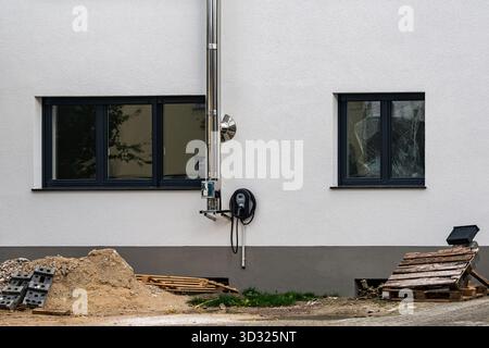 Ein neu erbautes Gebäude verfügt über zwei dunkle Fensterrahmen und ein Rohr, das sich entlang der Wand erstreckt. In der Nähe befinden sich Schmutzhaufen, Holzpaletten und Ziegelsteine Stockfoto