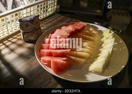 Gemütliches Stillleben auf weißem Teller mit frischen Fruchtscheiben. Gesunder Snack mit Wassermelonen-, Melonen- und Ananasdessert auf dunklem Holztisch in warmer Atmosphäre Stockfoto