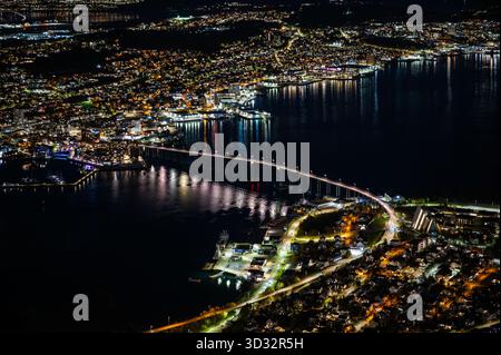 Tromso, Norwegen, atemberaubender nächtlicher Luftanblick auf eine pulsierende Stadt mit einer Brücke über ein ruhiges Gewässer, umgeben von beleuchteten Gebäuden und einem Bu Stockfoto
