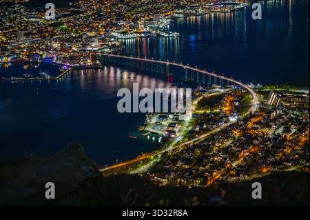 Tromso, Norwegen, atemberaubender nächtlicher Blick auf die pulsierende Stadt am Wasser, beleuchtet von Lichtern, die vom ruhigen Meer reflektiert werden, und einer langen Brücke Stockfoto