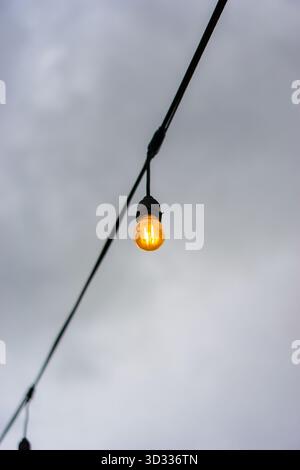 Minimalistisches Foto einer einzelnen hängenden Glühbirne, die warm auf einem Draht vor einem wolkigen grauen Hintergrund leuchtet. Stockfoto
