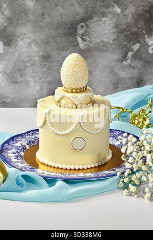Kleiner, eleganter osterkuchen mit weißer Schokolade, Kokosnussei, goldenen und weißen Perlen auf hellblauem Stoff Stockfoto