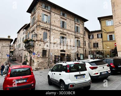 Assisi, Italien - 16. Oktober 2024: Charmante Steinarchitektur in Assisi, Italien, mit geparkten Autos, Fußgängern und Cafés. Stockfoto
