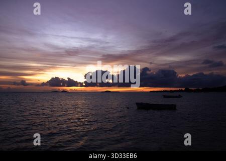 Sanfte tropische Dämmerung über weit entfernten Booten und schimmernde Reflexionen auf dem ruhigen Indischen Ozean in der Nähe von Praslin, Seychellen. Stockfoto