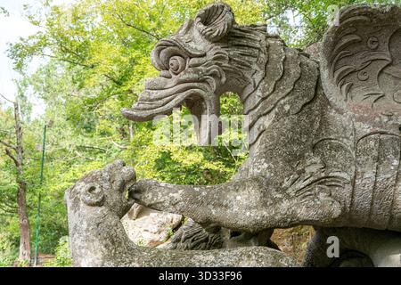 Italien – Bomarzo – Sacro Bosco – Drache kämpft gegen einen Löwen, aus nächster Nähe betrachtet die geflügelte Schlangenkreatur, die ihren Feind auf einem felsigen Sockel zwischen Bäumen packt Stockfoto