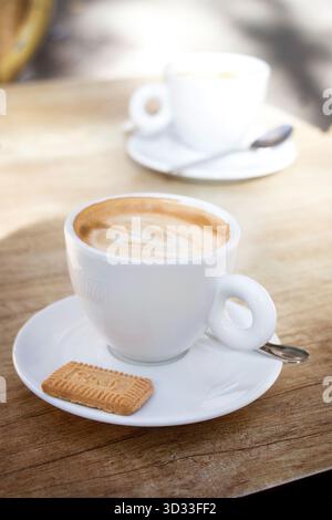 Nahaufnahme eines Cappuccinos, serviert in einer weißen Keramikschale mit Illy-Logo auf Holztisch mit einem Keks und einer anderen Tasse, die im Hintergrund verwischt ist palma mallorca Stockfoto