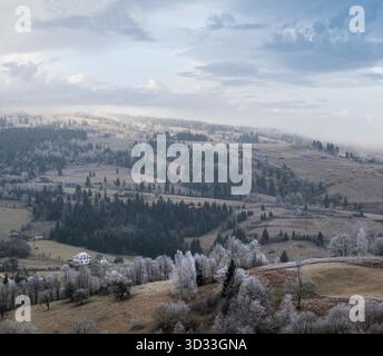 Der Winter kommt. Malerische Szene vor Sonnenaufgang über der Berglandschaft im Spätherbst mit Raureif auf Gräsern, Bäumen und Hängen. Friedliche Sonnenstrahlen vom bewölkten Himmel. Ukraine, Karpaten. Stockfoto