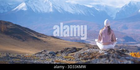 Eine Frau sitzt friedlich in Meditation auf einem Felsvorsprung, umgeben von atemberaubenden Bergen und farbenfrohen Herbstlandschaften. Sie umarmt die Natur Stockfoto