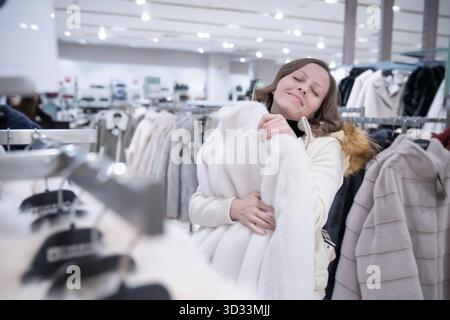 Eine weiße 30- bis 35-jährige Frau mit einem Lächeln umarmt in einer Modeboutique einen neuen Pelzmantel - sie träumt davon, zu Weihnachten einen neuen Pelzmantel zu bekommen. Stockfoto