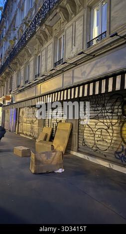 Pappkartons wurden an einer mit Graffiti bedeckten Wand in einer städtischen Straße entsorgt. Die geschlossene Ladenfront mit gestreiften Markisen unterstreicht die ruhige, frühe e Stockfoto