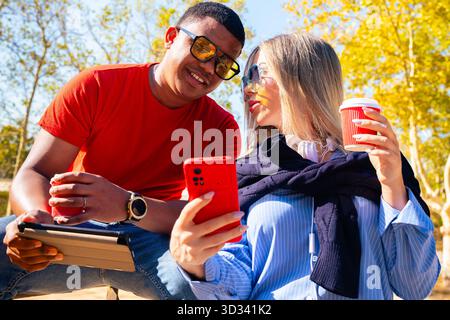 Lächelnde Freunde im Freien, die auf das Smartphone schauen und Kaffee trinken. Stockfoto