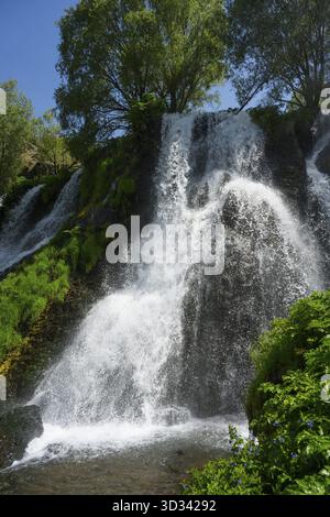 Ein Wasserfall fließt über Felsen, umgeben von Bäumen und grünem Laub unter einem blauen Himmel, Shaki Wasserfall, Syunik, Armenien Stockfoto