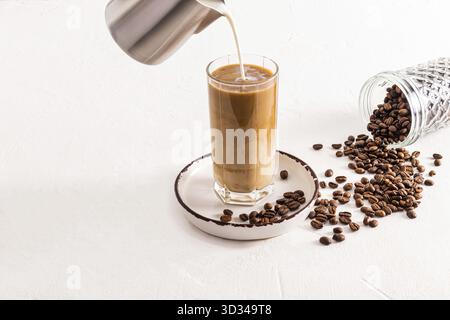 Ein Milchstrahl aus einer Metallkanne wird in ein hohes Glas eiskalten Kaffees gegossen. Dosen zur Aufbewahrung von Kaffee. Zubereitung eines köstlichen Getränks Stockfoto