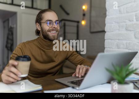 Fröhlicher bärtiger Mann freiberuflich in Brille, der zu Hause oder im Café mit Laptop arbeitet und Kaffee zum Mitnehmen trinkt Stockfoto