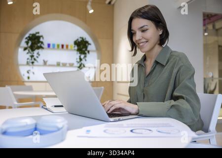 Lächelnd, zufriedene junge Geschäftsangestellte, die am Schreibtisch saß und am tragbaren Computer arbeitete Stockfoto