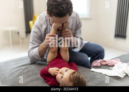 Mann, der mit der kleinen Tochter spielt, Vater, der sein Gesicht mit den Kinderfüßen versteckt, Fan mit dem Kleinkind zu Hause Stockfoto