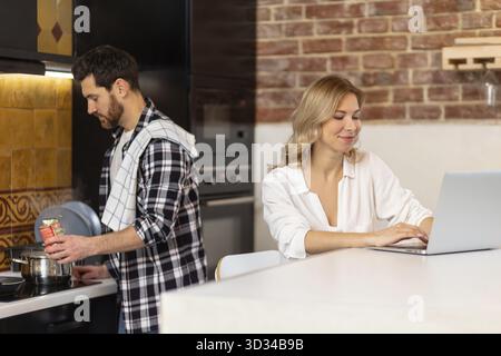 Frau, die am Computer in der Küche arbeitet, während ein Mann in kariertem Hemd das Abendessen vorbereitet Stockfoto