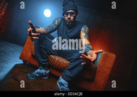 Junger Mann Breakdancer, der in einem großen alten Stuhlporträt sitzt. Tätowierung an den Händen Stockfoto