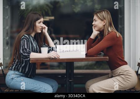 Frau hält weißes Blatt Papier beschriftet Wort Depression in der Hand. Zwei Frauen im Straßencafé. Versteckte Depression Konzept Stockfoto