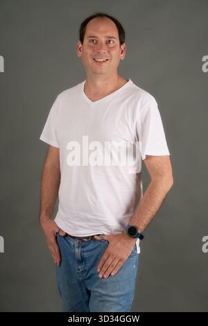 Ein lächelnder junger Mann in weißem T-Shirt mit V-Ausschnitt und Jeans posiert selbstbewusst vor einem neutralen grauen Studiohintergrund. Lässiger Stil, entspannte Attitüde, sauber Stockfoto