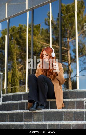 Eine junge Frau mit fließenden roten Haaren sitzt draußen und trägt weiße Kopfhörer. Sie hält ein Smartphone und schaut nachdenklich auf ihre Umgebung, enjoyi Stockfoto