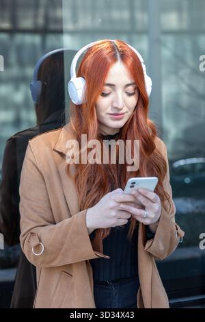 Eine junge Frau mit langen roten Haaren genießt Musik auf ihrem Telefon, während sie elegante Kopfhörer trägt. Sie steht vor einem Glasgebäude und lächelt den C an Stockfoto