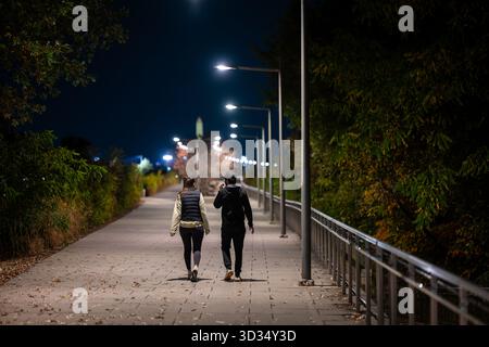 Menschen auf dem Pfad Long Bridge Park Arlington / ARLINGTON, VIRGINIA – zwei Personen spazieren nachts auf einem gepflasterten Pfad im Long Bridge Park, der von modernen Straßenlaternen beleuchtet wird. Dieser urbane Park in Arlington ist bekannt für seine Freizeiteinrichtungen und die malerische Aussicht mit weitläufigen Grünflächen, Sportplätzen und Blick auf Washington D.C. und den Potomac River. Der Pfad ist von Bäumen gesäumt, von denen einige herbstliches Laub zeigen, und gefallene Blätter sind auf dem Boden verstreut, was eine entscheidende Verbindung zum Mount Vernon Trail darstellt. Stockfoto