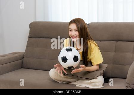 Junge glückliche Frau zu Hause, die sportliche Aktivitäten genießt, bequem auf einem gemütlichen Sofa sitzt und Fußball hält Stockfoto