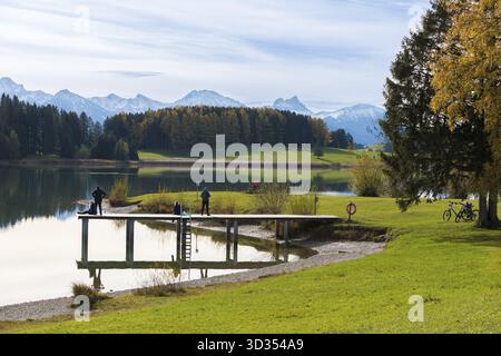 Die Leute laufen entlang des Stegs mit Blick auf den Forggensee und die umliegenden Berge, Herbstbäume im Hintergrund, bei Rosshaupten, Ostallgäu, alles Stockfoto