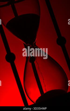 Nahaufnahme einer Sanduhr mit Sand fließend, Silhouetten gegen leuchtendes Rot. Ein dramatisches Symbol für Zeit, Dringlichkeit und philosophische Reflexion. Stockfoto