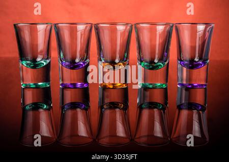 Fünf Klarsichtgläser mit farbigen Sockeln auf rot-orangefarbenem Hintergrund. Reflexionen auf glänzender Oberfläche verbessern die Symmetrie und die lebendige visuelle Wirkung. Stockfoto