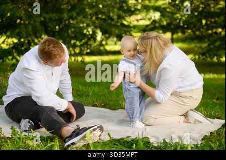 Fröhlicher Familienmoment im Park: Das Baby macht mit Unterstützung der Eltern erste zaghafte Schritte, und glückliche Mutter küsst das süße Baby. Familie genießt Zeit im Freien Stockfoto