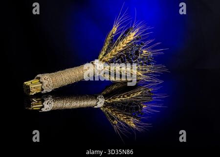 Dekoratives Bündel aus Weizen und getrocknetem Lavendel, mit Spitze gebunden, auf eine schwarze reflektierende Oberfläche gelegt. Eine rustikale und elegante Komposition. Stockfoto