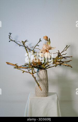 Das einzigartige Blumenarrangement zeigt zarte Blüten und Zweige in einer strukturierten Vase auf einem Sockel und schafft so ein elegantes und doch natürliches Herzstück Stockfoto