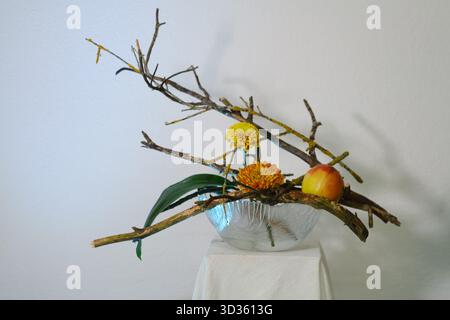 Ein einzigartiges Blumenarrangement mit Zweigen, hellgelben Blüten und einem Pfirsich, präsentiert auf einem minimalistischen Sockel vor weißem Hintergrund. Die DE Stockfoto