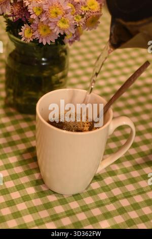Heißes Wasser wird in eine weiße Tasse mit Kaffee gegossen. Im Hintergrund eine grüne karierte Tischdecke und ein Glas mit rosa Chrysanthemen. Oben Stockfoto
