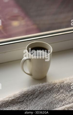 Weiße Tasse mit Kaffee auf einer Fensterbank, graue Decke in der Nähe, verschwommenes rotes Dach draußen. Stockfoto