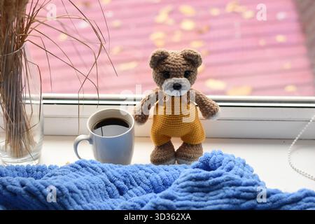 Gestrickter Teddybär mit Kaffeetasse und Vase mit trockenen Stielen auf blauer Decke, gemütliche Heimszene. Stockfoto