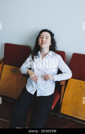 Eine junge Frau mit lockigem Haar, in einem schicken weißen Hemd und einer eleganten schwarzen Hose, entspannt sich auf Vintage-Stühlen und verkörpert wahren Komfort und Freizeit mit einem Ser Stockfoto