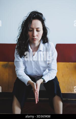 Eine Frau in einem weißen Hemd sitzt auf einer Holzbank, ihre Hände sind zusammengebunden. Sie scheint in Gedanken verloren zu sein, umgeben von einer ruhigen Atmosphäre, die einlädt Stockfoto