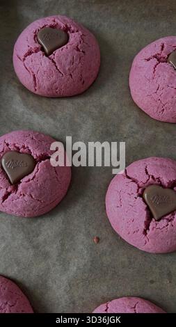 Vertikale Nahaufnahme mit hausgemachtem rosa Samtkekse oder roten Samtkeksen mit einem geschmolzenen Milka-Schokoladenherz, gebacken auf einem Pergamentpapier Stockfoto