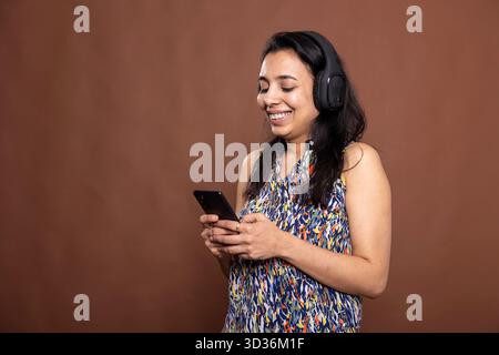 Glückliche indianerin, die Musik auf schwarzen kabellosen Kopfhörern und SMS auf ihrem Smartphone hört. Ein hübsches weibliches Model lächelt auf den Bildschirm des Geräts, während es ein lässiges Outfit trägt, isoliert auf dem Studiohintergrund. Stockfoto