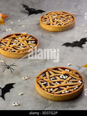 Drei Halloween-Torten sitzen auf einer grauen Oberfläche, verziert mit Spinnennetzen und Geisterformen. Schwarze Fledermäuse und Herbstblätter verleihen dem Löffel eine festliche Note Stockfoto