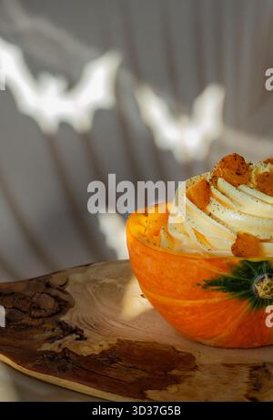 Ein kreativ gestaltetes Kürbisdessert sitzt in einer geschnitzten Kürbisschale, garniert mit Toppings, auf einem rustikalen Holzbrett. Natürliches Licht verstärkt den Krieg Stockfoto