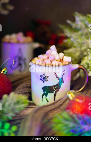 Ein gemütliches Getränk gefüllt mit Marshmallows sitzt in einem festlichen Becher, der mit einem Rentier geschmückt ist. Es ist umgeben von farbenfrohen Ornamenten und Weihnachtsgrün, Crea Stockfoto